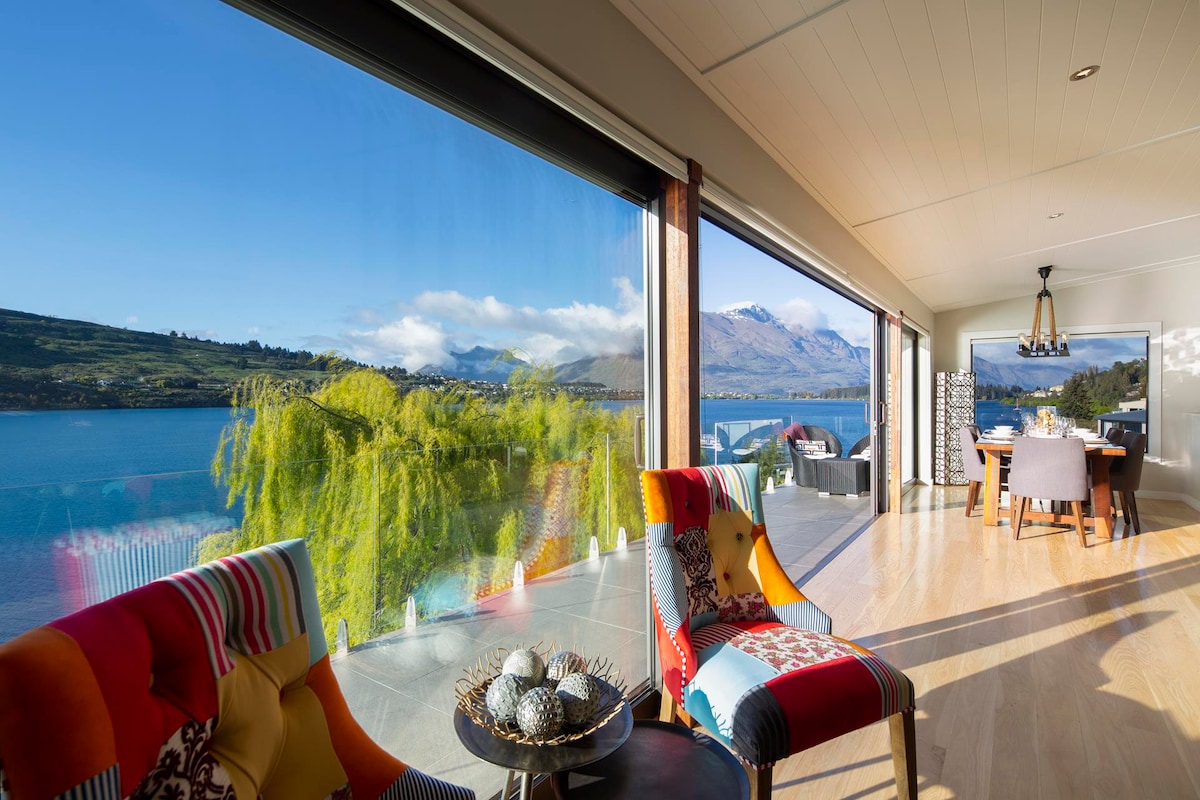 A spacious living area is highlighted by large floor-to-ceiling windows that provide unobstructed views of the lake and mountains. Colorful chairs frame a stylish coffee table, creating a welcoming space for relaxation. The open layout leads to a dining area, enhancing the connection to the outdoor scenery.