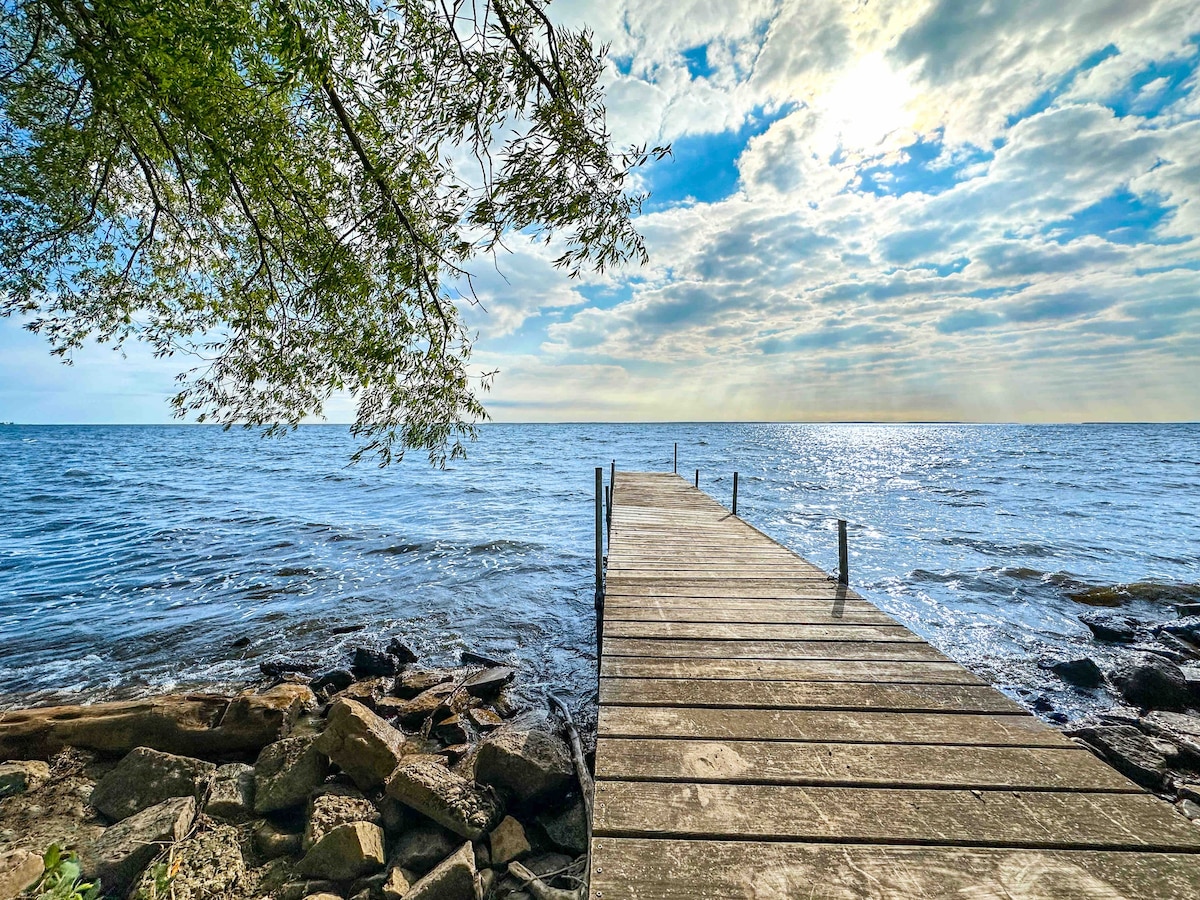 A wooden dock extends into the calm waters of Green Bay, framed by a leafy tree branch. Rocks line the shoreline, while soft clouds and sunlight reflect off the water's surface, creating a serene outdoor setting.