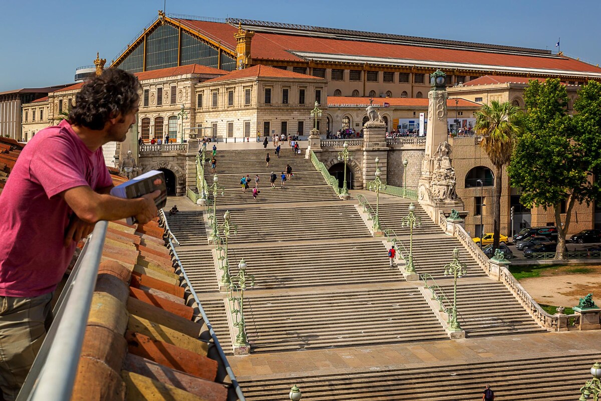 The image captures the majestic staircase of the Saint-Charles railway station in Marseille, leading up from the foreground. Visitors can be seen ascending the steps, surrounded by detailed architectural features and greenery, while the bright blue sky overhead provides a vibrant backdrop.