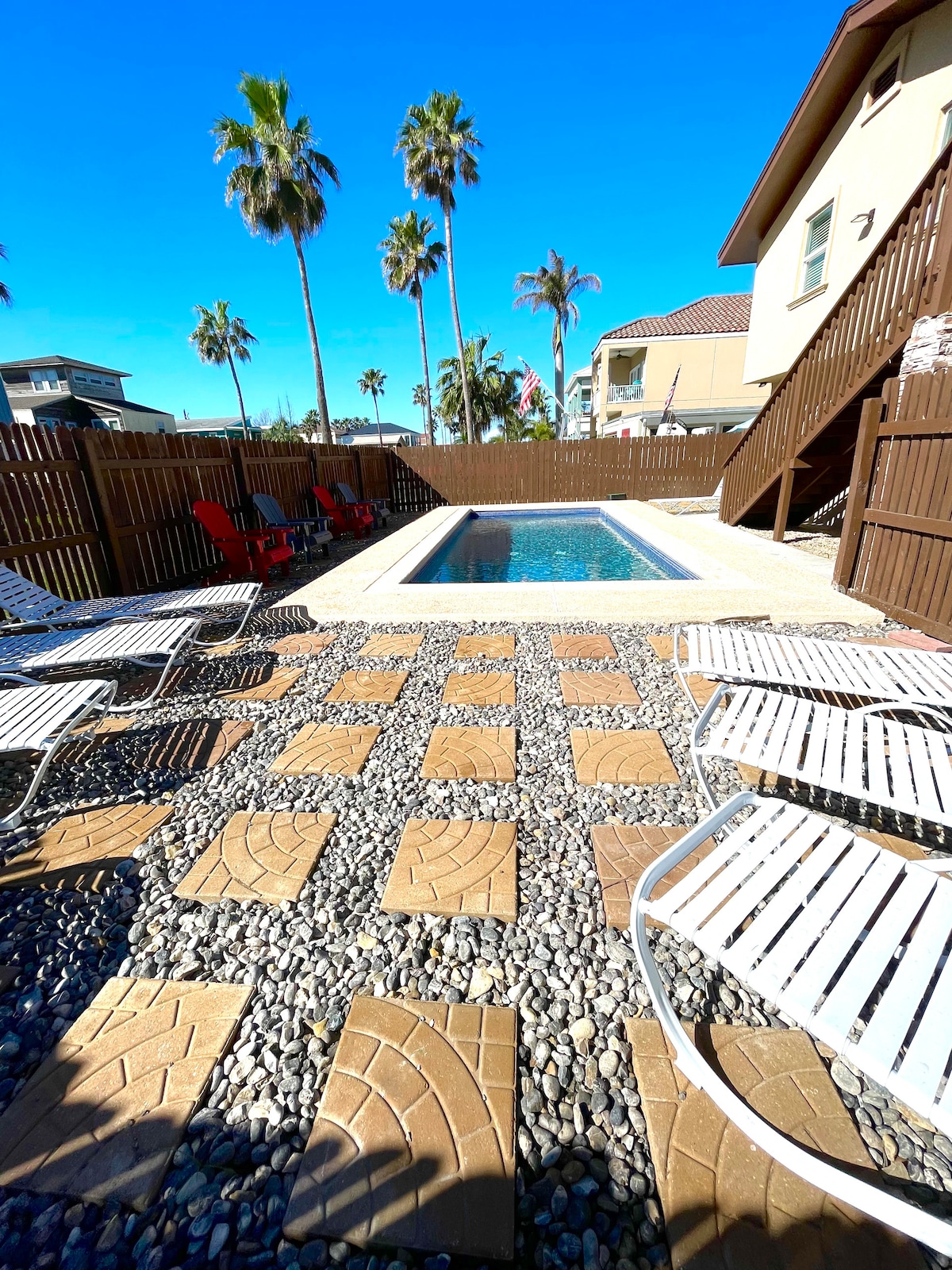 A sunlit outdoor pool area is surrounded by a mix of textured stone and concrete pathways. Lounge chairs are placed for relaxation under the clear blue sky, while tall palm trees offer a tropical ambiance. A wooden fence provides privacy to the space.