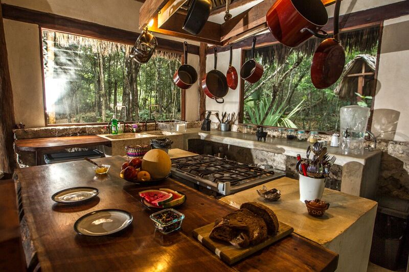 A spacious kitchen features a large island with wooden countertops and an array of cooking utensils. Hanging pots reflect natural light from large windows, showcasing views of the surrounding jungle. Plates and fresh fruits are prominently displayed, emphasizing a connection to nature and culinary experiences.
