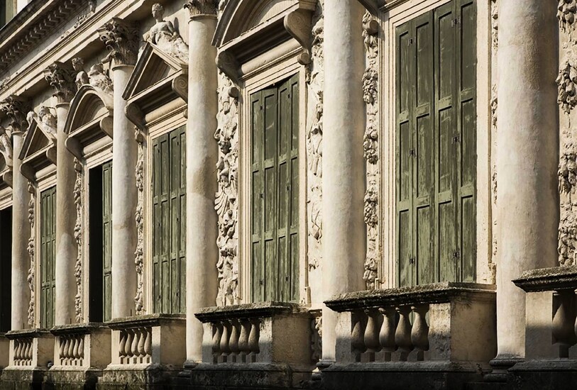 A historic building facade is highlighted, featuring large, ornate windows adorned with decorative molding. The wooden shutters, painted in a muted green hue, are partially closed, complementing the ancient stone structure's architectural details.