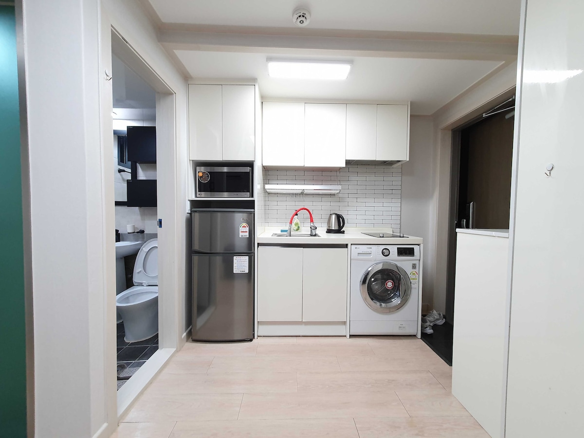 The kitchen area features modern appliances including a refrigerator, stove, and washing machine. Light-colored cabinetry and white tile backsplash create a clean look. A doorway leads to a bathroom, visible in the left portion of the image.