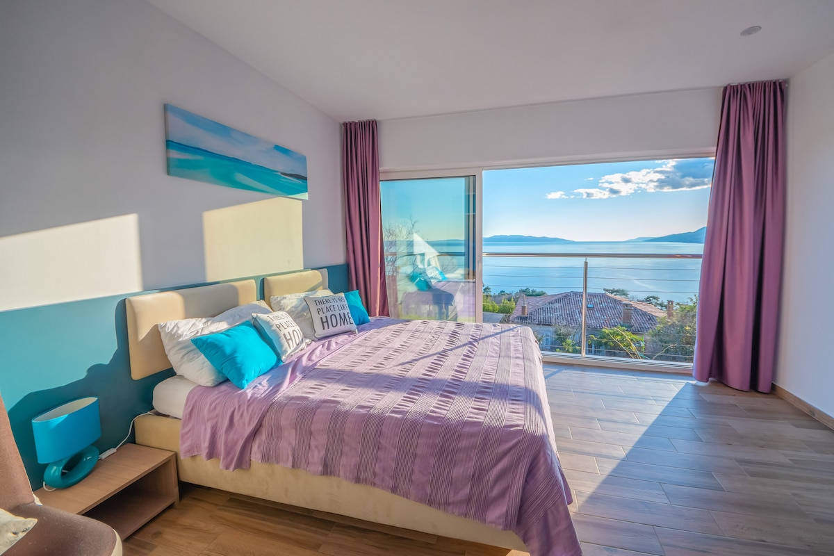 A spacious sleeping area features a large bed adorned with patterned pillows and a soft throw. Floor-to-ceiling windows offer a panoramic view of the sea, while light drapes gently frame the windows, allowing natural light to fill the room.
