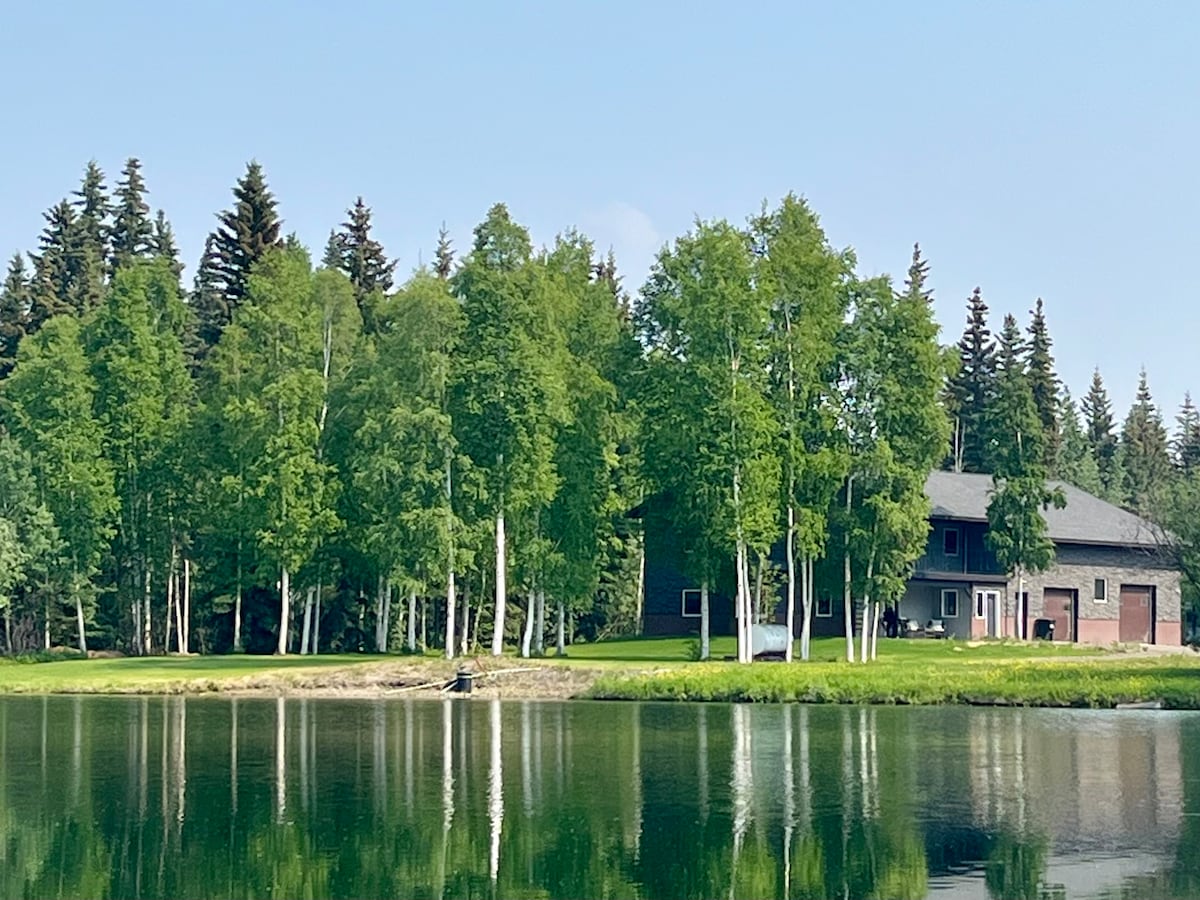 A waterfront view reveals a rustic hangar home nestled among lush green trees, reflecting on the calm water surface. The structure features a mix of natural wood and modern exterior design, complemented by a surrounding sandy beach area.