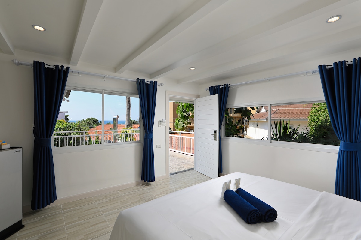A spacious room features large windows that provide natural light and a view of the surrounding greenery. Blue curtains frame the windows, and a simple bed is made with white linens and neatly rolled towels. The area is clean and inviting, with a hint of outdoor tranquility.