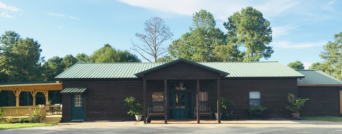 A rustic wooden structure features a green metal roof and a welcoming front entrance. Flanking the entrance, benches are placed for seating, while manicured shrubs and trees create a natural backdrop. A spacious outdoor deck is visible on one side, enhancing the overall charm.