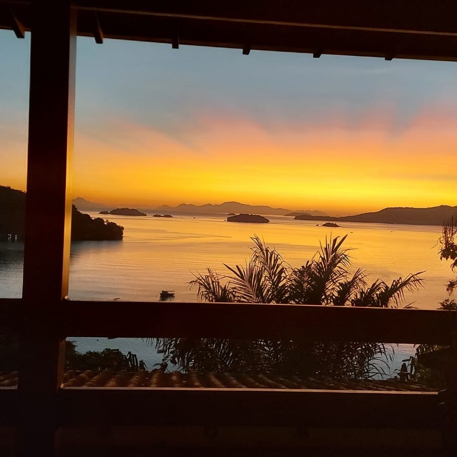 A vibrant sunset casts warm hues over the tranquil sea, framed by wooden beams. Silhouettes of distant islands are visible against the horizon, with lush greenery enhancing the serene landscape. The scene captures a peaceful moment in nature.