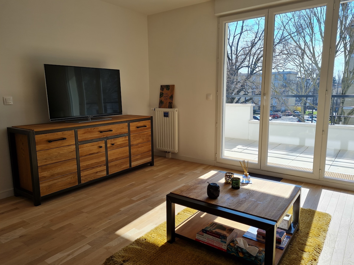 A living area features a wooden console table with a flat-screen television above. A wooden coffee table rests on a textured yellow rug. Large glass doors allow ample natural light and provide views of the balcony and outdoor trees.