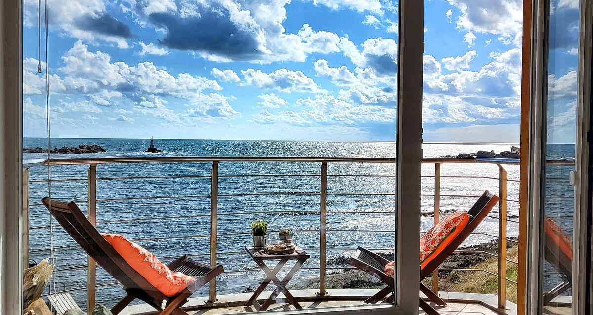 A balcony view showcases two wooden lounge chairs facing the open sea, with sunlight reflecting on the water. A small table holds decorative elements. The sky features scattered clouds, enhancing the overall tranquil atmosphere.