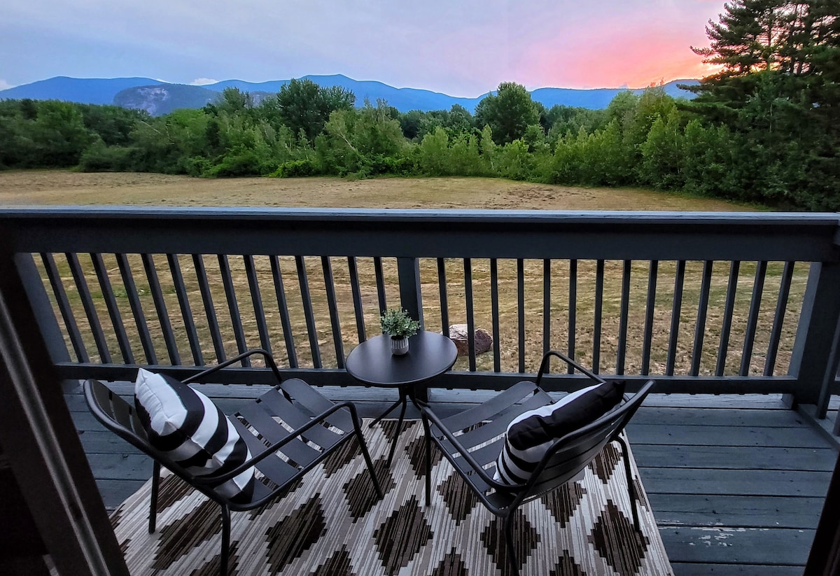 A serene outdoor balcony is furnished with two stylish chairs and a small table, creating an inviting space for relaxation. A striped area rug adds texture, while the expansive view of rolling hills and greenery captures the beauty of the surrounding landscape under a colorful sunset sky.