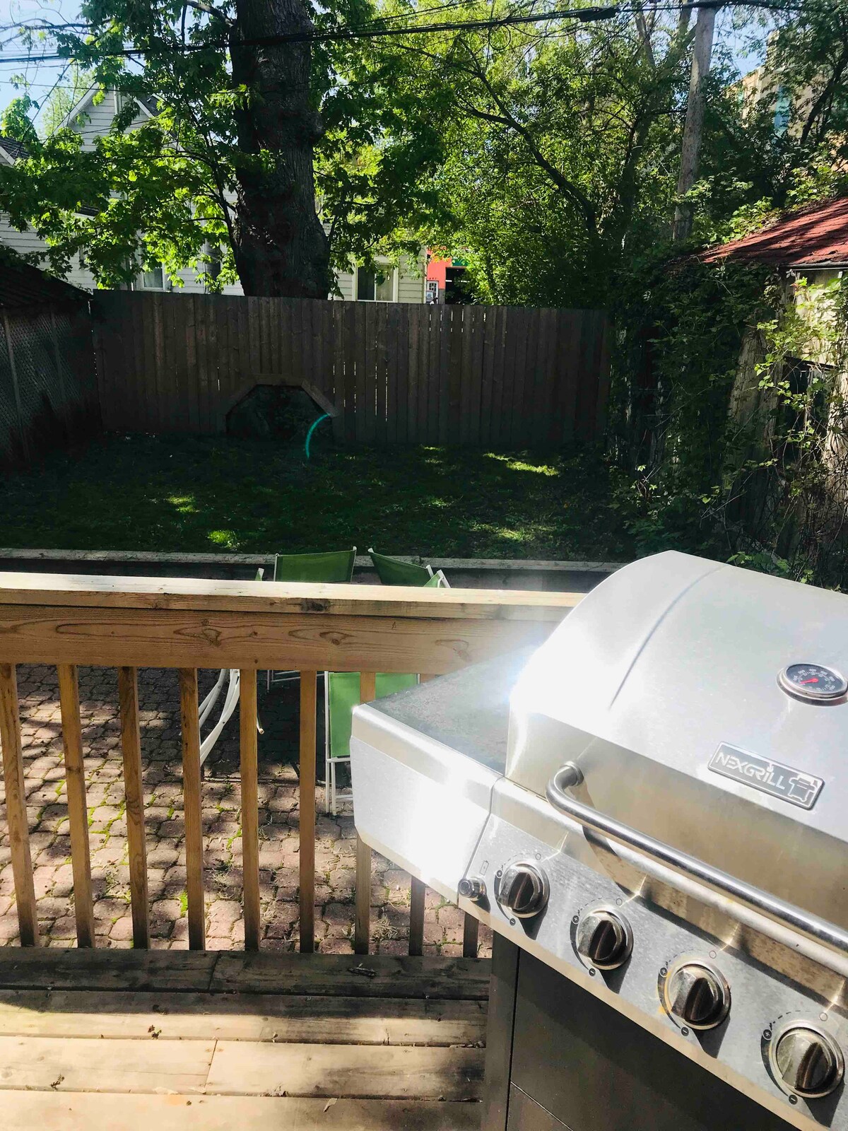 A stainless steel grill is positioned on a wooden deck, overlooking a grassy backyard surrounded by a wooden fence. Greenery from trees and shrubs creates a natural setting, while an inviting atmosphere is suggested by the open space.