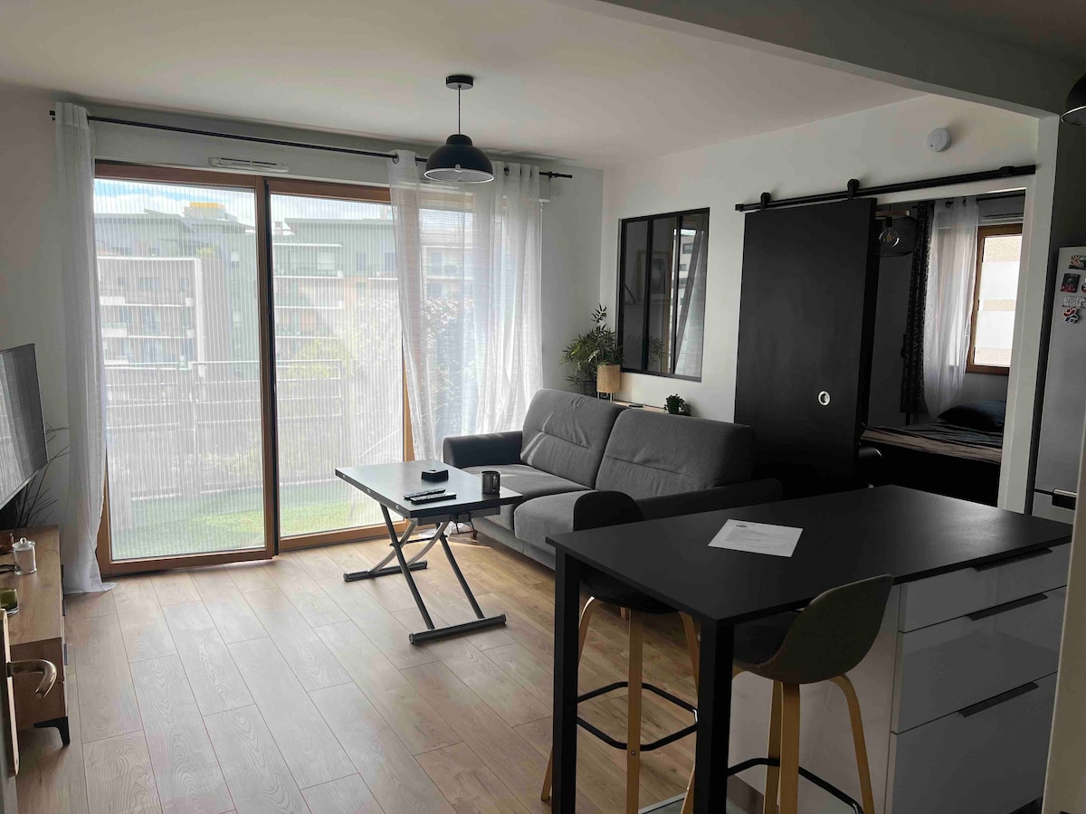 A bright living area is observed, featuring large glass doors that open to a private outdoor space. A light gray sofa is positioned beside a small table. A dark sliding door leads to a sleeping area. Light wood flooring enhances the natural light in the room.
