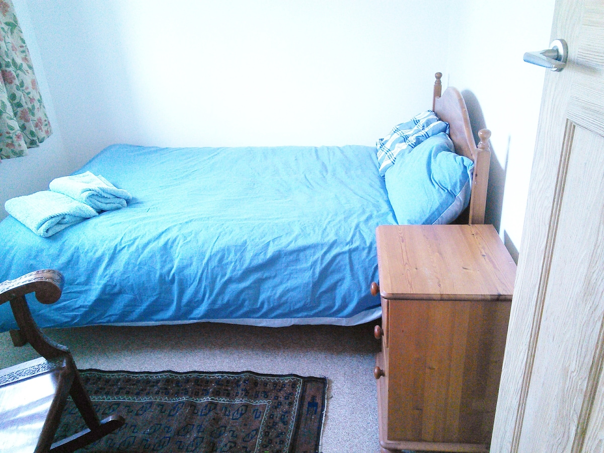 A simple bedroom features a single bed dressed in a light blue cover, complemented by a small wooden bedside table. A woven rug adorns the floor, and a rocking chair is positioned near the door. Natural light filters through floral curtains.