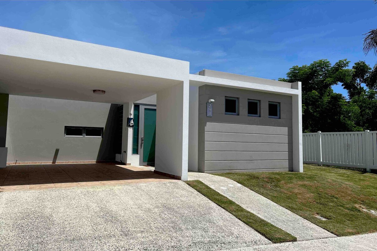 A modern, single-story home is showcased with a carport and a welcoming entrance. The exterior features light-colored walls, large windows, and a neatly manicured lawn. A pathway leads toward the front door, bordered by well-maintained greenery.