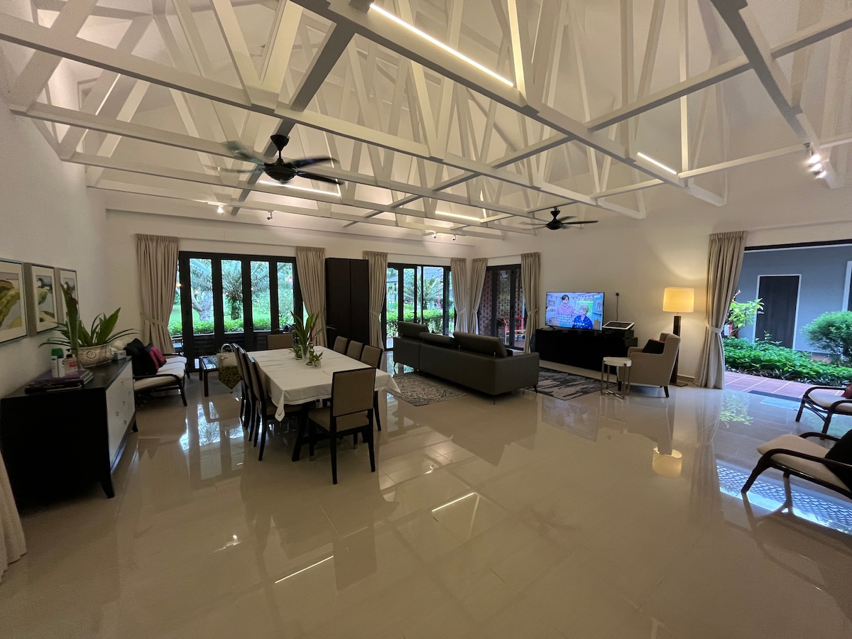 The spacious living room features a large sofa facing a television, with a dining table nearby. Large windows invite natural light and provide views of the surrounding greenery. The modern decor is complemented by ceiling fans and light fixtures, enhancing the room's open feel.