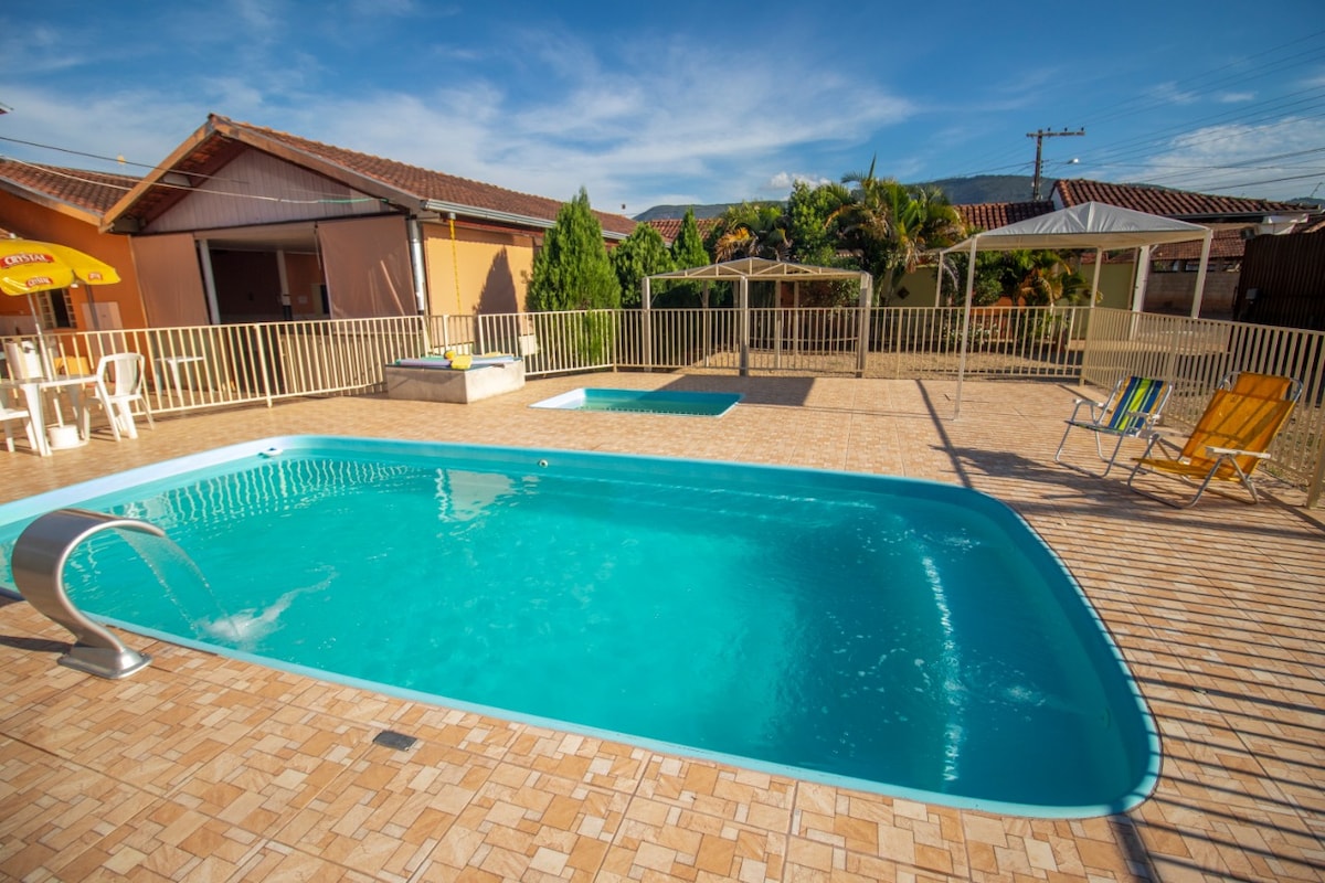 A large swimming pool is pictured, featuring clear blue water and a gentle fountain. Surrounding the pool area are two sun loungers, a small shaded gazebo, and a second smaller pool. The patio is lined with tiled flooring, creating an inviting outdoor space.
