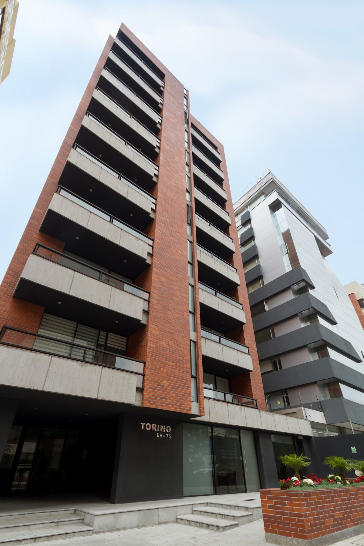 A modern apartment building features multiple floors with a striking brick facade and large balconies. A clean entrance area is visible, with a minimalist design complemented by greenery. The structure's contemporary architecture reflects a blend of style and functionality in a well-maintained urban setting.
