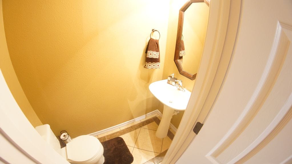 A compact bathroom features warm yellow walls and a curved mirror above a pedestal sink. A tasteful towel rack holds two hand towels, and tiled flooring adds a polished look. A neutral mat lies in front of the toilet, creating a cohesive style.