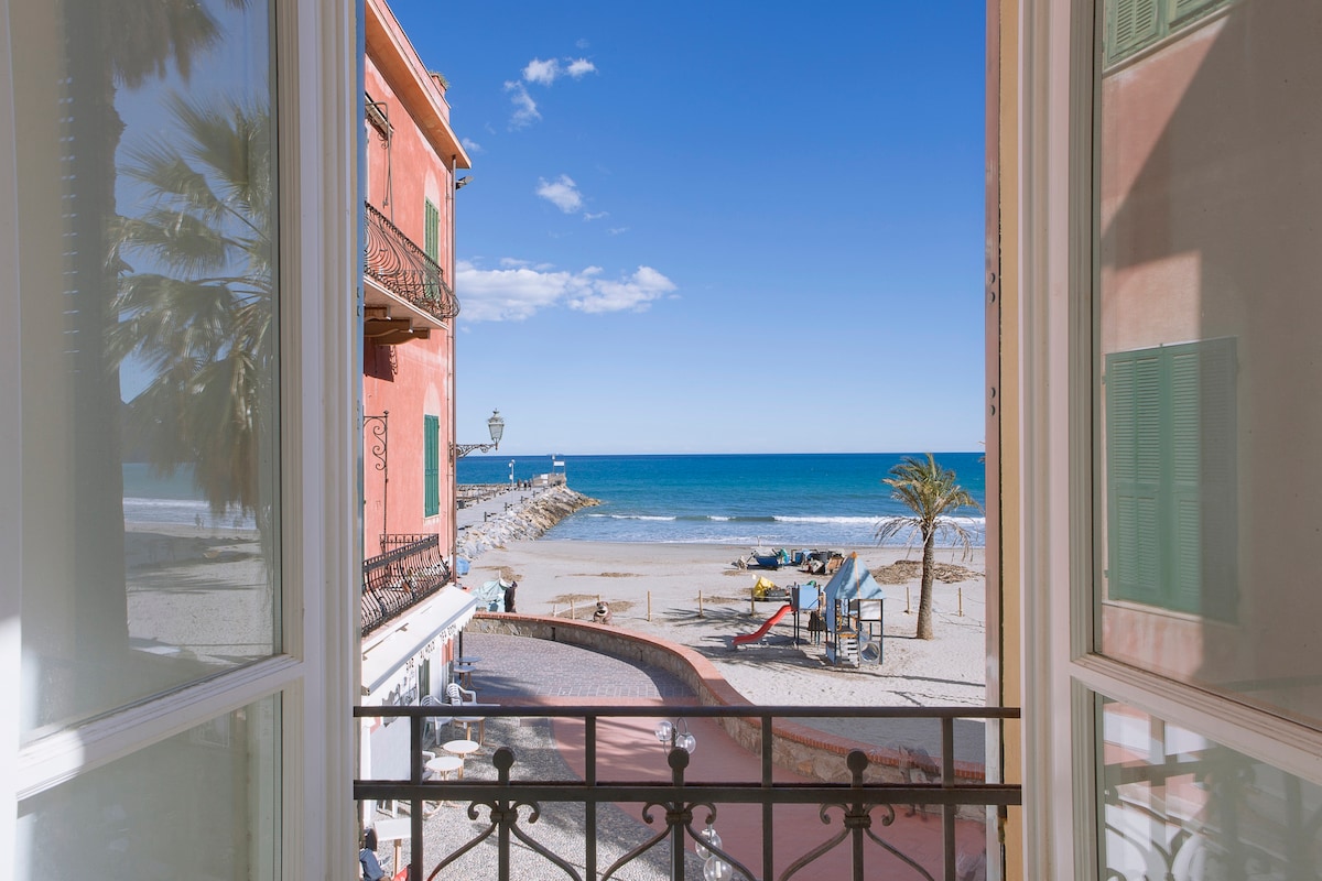 A view from the window reveals a sunny beachfront scene, with clear blue waters and a sandy shore. Palm trees and colorful buildings frame the vista, while a promenade is visible, along with parked vehicles and recreational equipment.