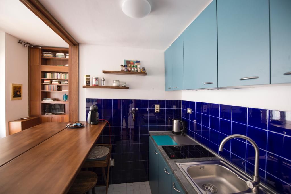 The kitchen features blue tile walls and modern cabinetry in a vibrant shade. A spacious wooden counter with stools offers a casual dining area. Open shelving displays various kitchen essentials, while appliances such as a kettle and coffee maker are visible upon the counter.