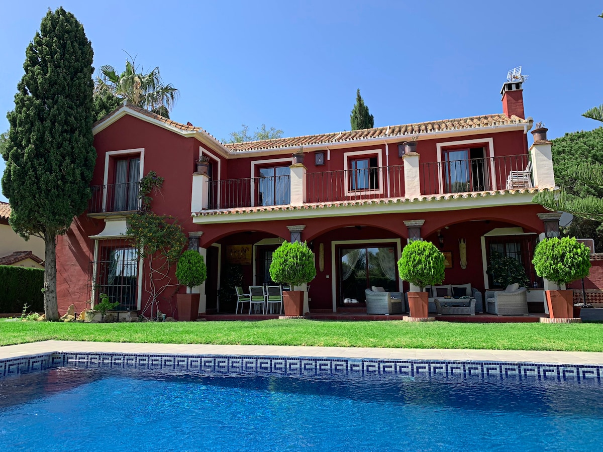 The exterior of the villa is displayed, featuring a two-story red structure with multiple windows and balconies. A well-maintained lawn surrounds a blue swimming pool, reflecting the clear sky. Tasteful greenery, including potted shrubs, complements the elegant outdoor setting.
