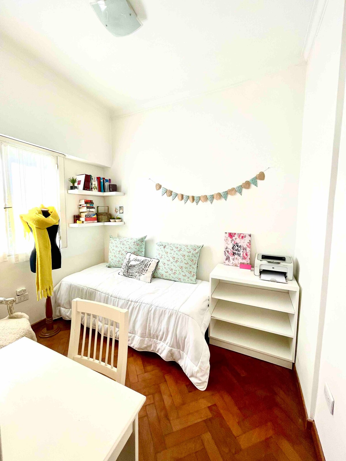 A bright and tranquil bedroom features a spacious daybed adorned with decorative pillows. A desk and chair provide a workspace, while a bookshelf displays an array of books. Natural light enters through a nearby window, enhancing the calm atmosphere.