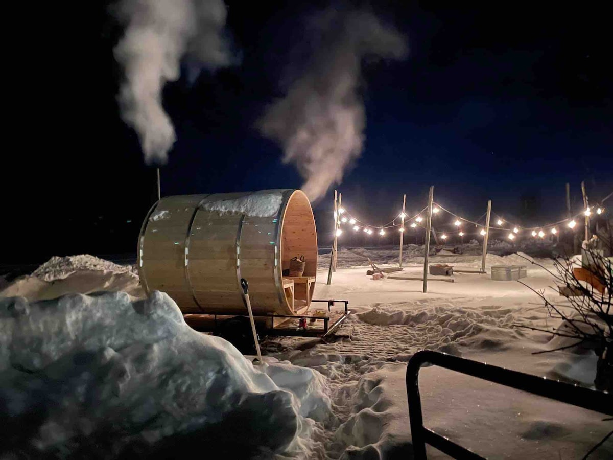 A wooden sauna is featured in a snowy landscape at night. Steam rises from the sauna, creating a serene atmosphere. Softly glowing string lights are strung overhead, illuminating the surrounding snow and creating a peaceful retreat setting. The scene captures the tranquility of a winter night.