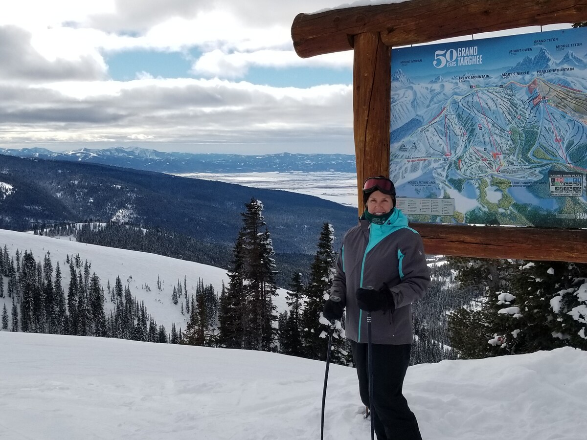 A snow-covered mountaintop is visible, showcasing a ski area map sign mounted on a rustic wooden post. Majestic mountain ranges stretch in the background under a partly cloudy sky, while tall evergreen trees frame the scene, emphasizing the beautiful winter landscape.