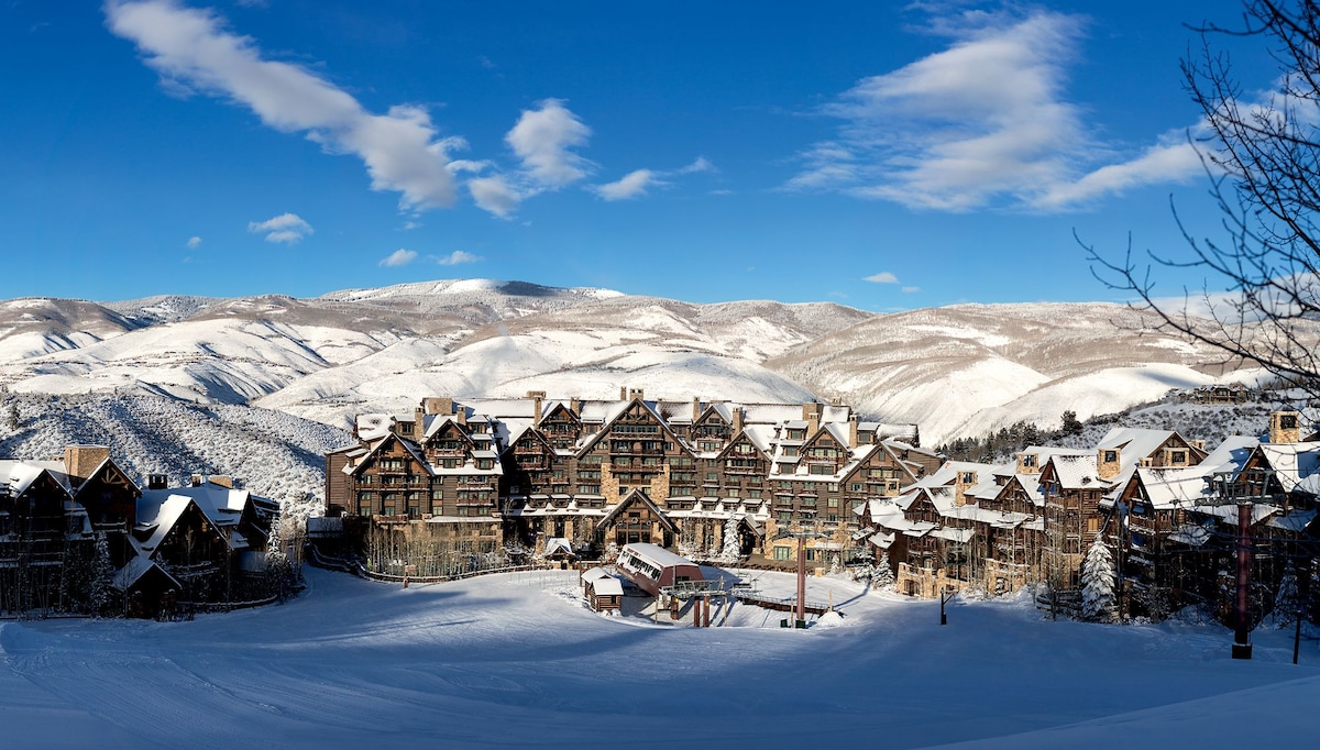 The expansive resort is set against a stunning winter landscape, surrounded by snow-covered mountains. The majestic building features a blend of rustic architecture, large windows, and balconies, with clear skies above adding to the serene atmosphere.