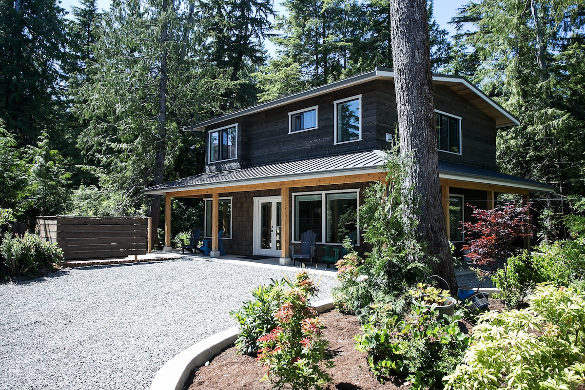 The exterior of the Sea Rogue Guest House is surrounded by lush trees, with a spacious gravel driveway leading up to the home. The two-story structure features a modern design, large windows, and a covered porch with seating, inviting a connection with nature.