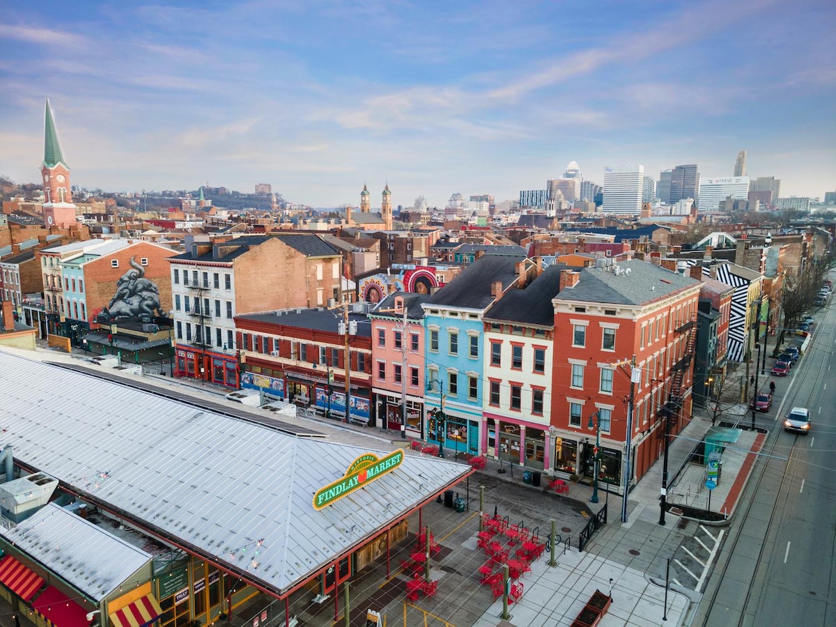 An elevated view captures the vibrant streetscape of Findlay Market, showcasing colorful historic buildings lining the street. The market's canopy and red seating area are visible, while the skyline of Cincinnati and a spire from a nearby church create a dynamic urban background.