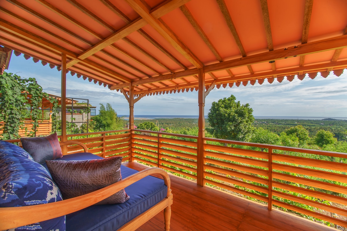 A wooden pavilion features a cozy seating area with a deep blue cushion. Vistas of lush greenery and the horizon are framed by slatted wooden railings under a terracotta roof.