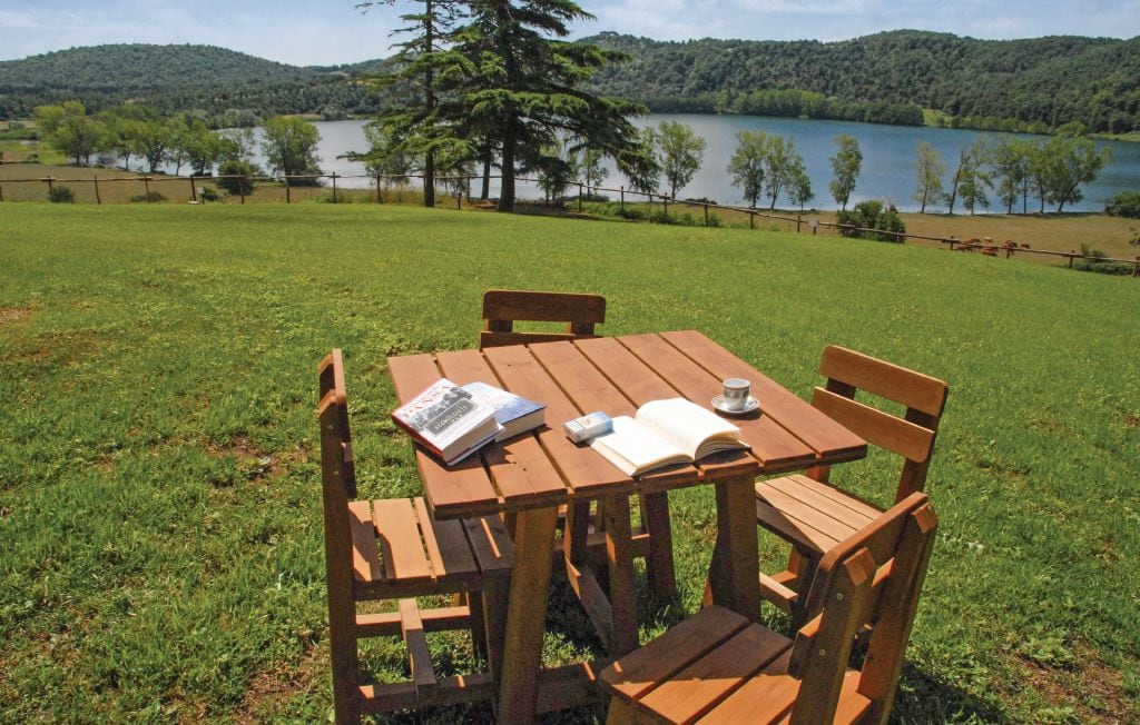 An outdoor wooden table with four chairs is set on a lush green lawn overlooking a serene lake. A book and a coffee cup are placed on the table, while trees and rolling hills frame the scenic view in the background.