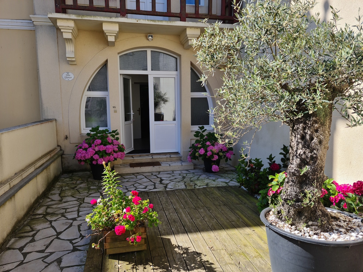 An inviting entrance features a stone pathway leading to a large front door, framed by vibrant pink hydrangeas and greenery. A mature olive tree stands prominently beside a wooden deck, which is adorned with colorful planters showcasing blooming flowers.