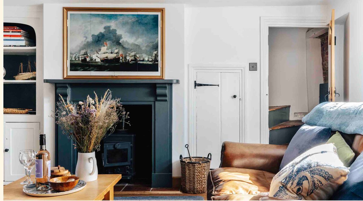 The living room features a leather sofa adorned with a decorative pillow, complemented by a wooden coffee table with a wine bottle and glasses. A floral arrangement is positioned in a vase atop the table. A cast iron fireplace is highlighted beneath a large painting, while a staircase is visible in the background.