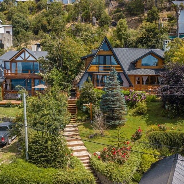 A charming house is situated on a hillside, surrounded by greenery and colorful flowers. A pathway leads to the entrance, flanked by trees and well-maintained gardens. Multiple outdoor seating areas are visible, providing spaces for relaxation and enjoyment of the serene environment.