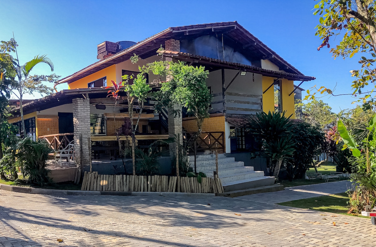 The exterior of a large, multi-level home is surrounded by greenery. A staircase leads to a welcoming entrance, with wooden structures and vibrant yellow walls visible. Various plants and trees enhance the natural setting, providing a serene atmosphere.