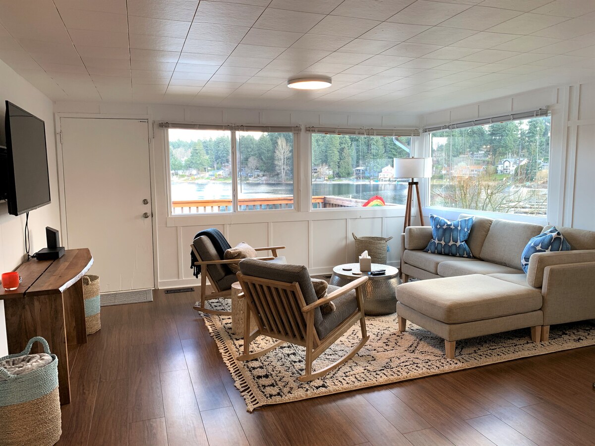 A bright living area features large windows overlooking the waterfront, allowing for ample natural light. Comfortable seating is provided by a spacious sectional couch and two armchairs. A patterned area rug adds warmth to the wooden floors, while a minimalistic coffee table offers a central gathering space.