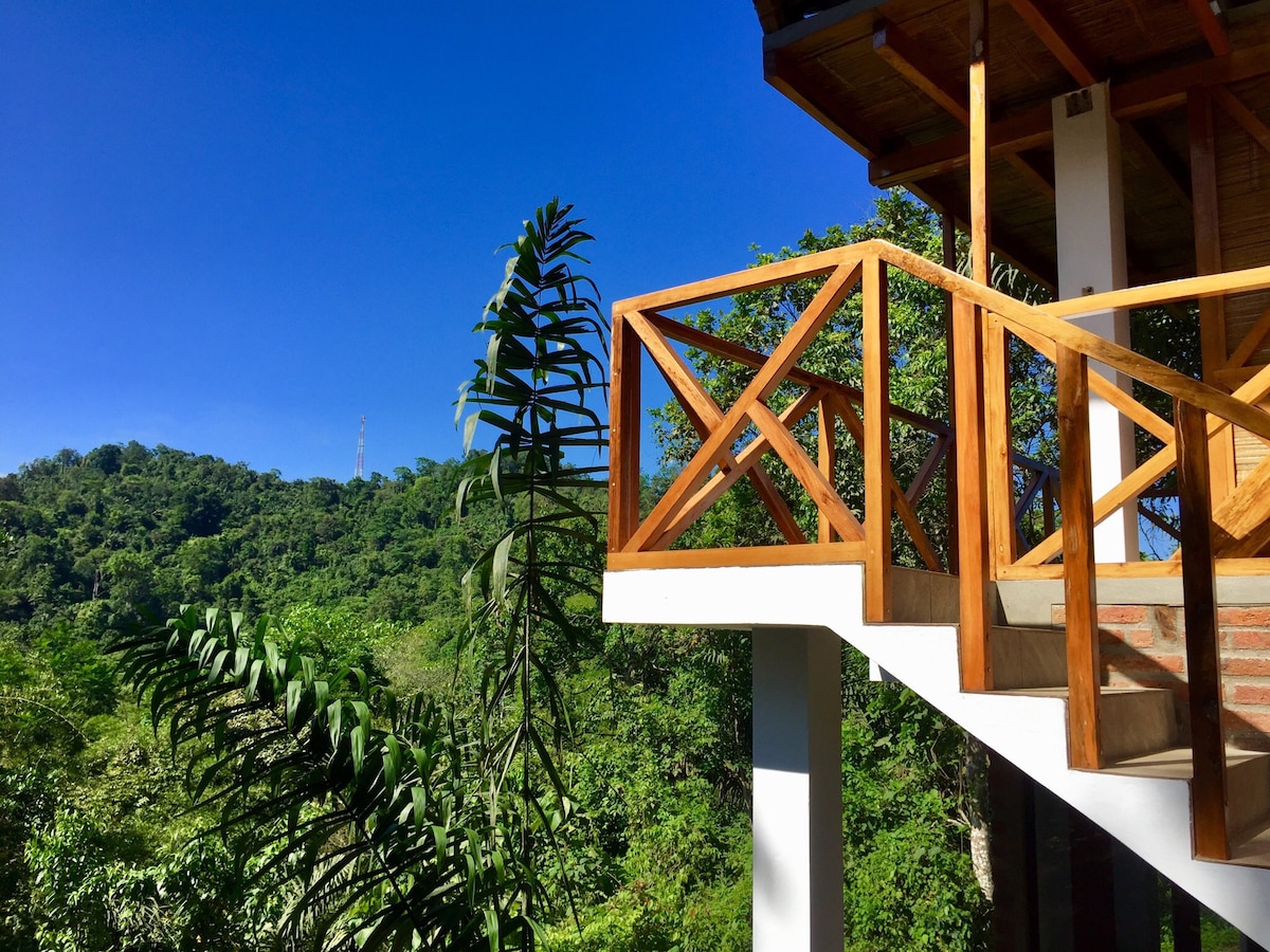 A wooden balcony railing offers views of the lush green hillside under a clear blue sky. The vibrant foliage creates a serene backdrop, enhancing the connection to nature. The structure's architectural details showcase a blend of natural materials with modern design elements.
