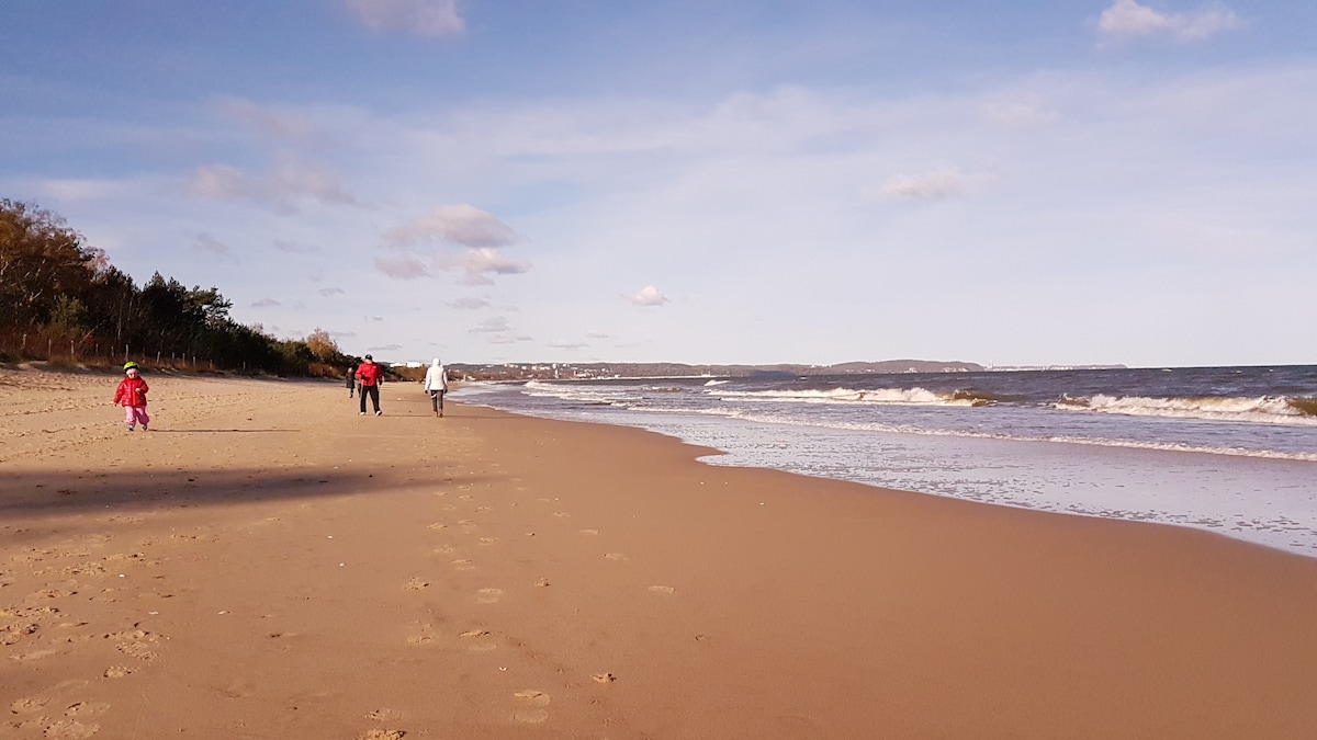 A sandy beach stretches along the shoreline, with gentle waves lapping at the coast. In the distance, three figures are seen walking along the water's edge, while a child in bright clothing plays nearby. The sky is partly cloudy, creating a serene coastal ambiance.