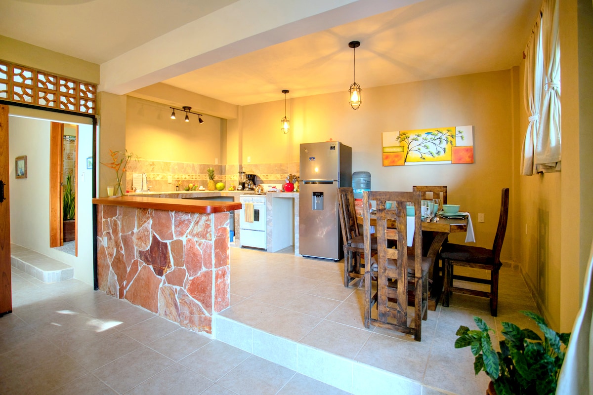 An open kitchen and dining area feature natural stone accents and wooden furniture. A large refrigerator and stove are visible, with countertop space for meal preparation. A dining table with chairs is positioned near a window, bathing the space in soft natural light.