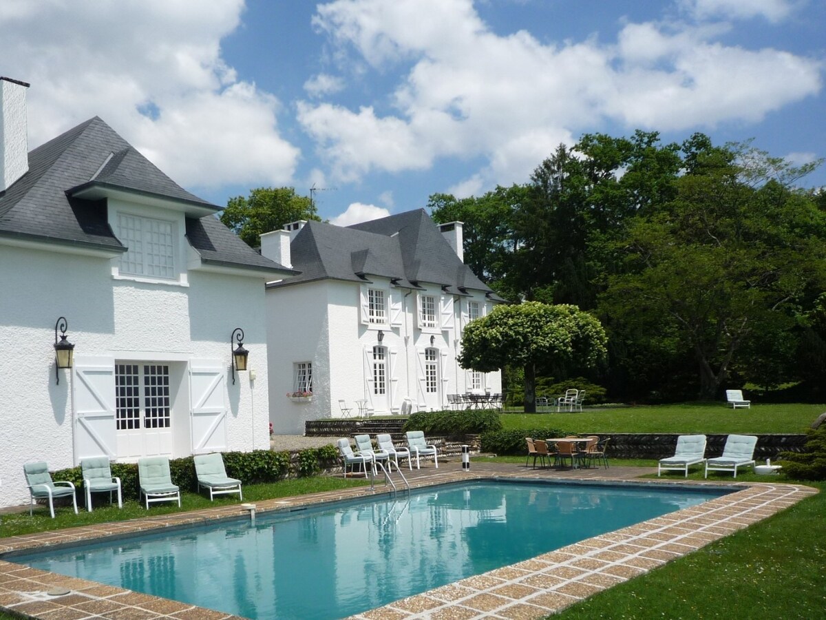An outdoor space features a serene pool surrounded by lounge chairs, with the white exterior of the manor house visible in the background. Lush green trees and well-manicured lawns create a calming atmosphere, while clouds drift across the blue sky.