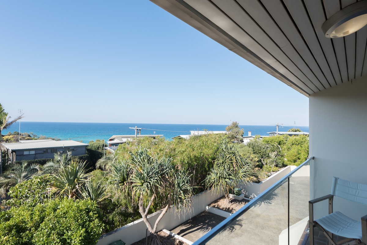 A scenic view of the ocean is presented from the balcony, with expansive blue waters visible in the distance. Lush greenery and coastal vegetation are seen in the foreground, complemented by clear skies above.