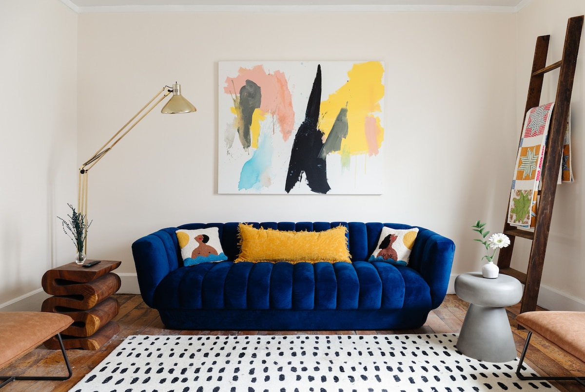 A vibrant living room features a deep blue velvet sofa adorned with colorful pillows. Above the sofa, a large abstract painting adds visual interest. A round table with a simple design stands nearby, accompanied by two light-colored chairs and a decorative ladder displaying a small artwork.