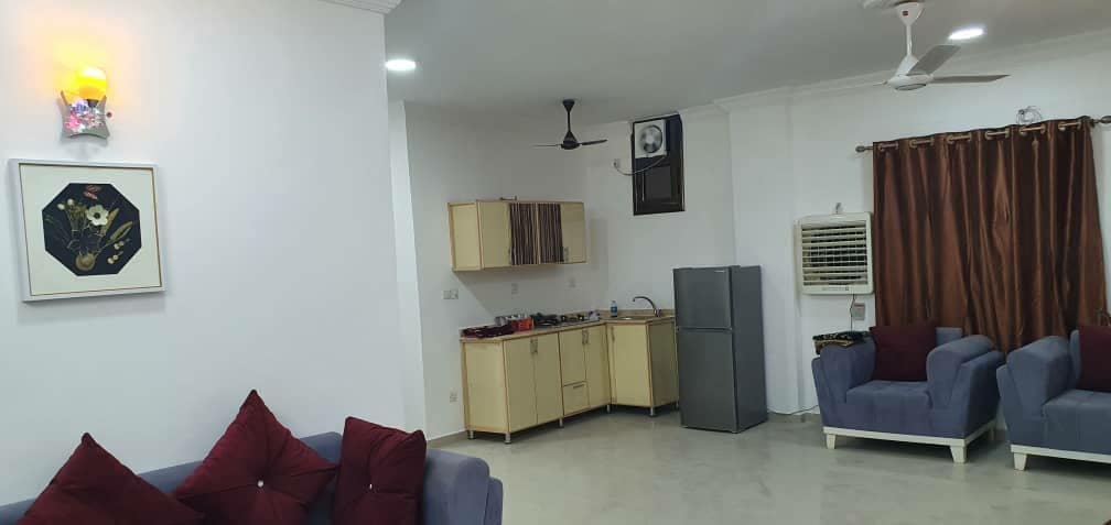 A spacious living area is featured, showcasing a modern kitchen with light cabinetry and essential appliances, including a gas cooker and refrigerator. Two comfortable blue sofas are arranged with decorative cushions. A large window is adorned with dark curtains, allowing natural light to fill the space.