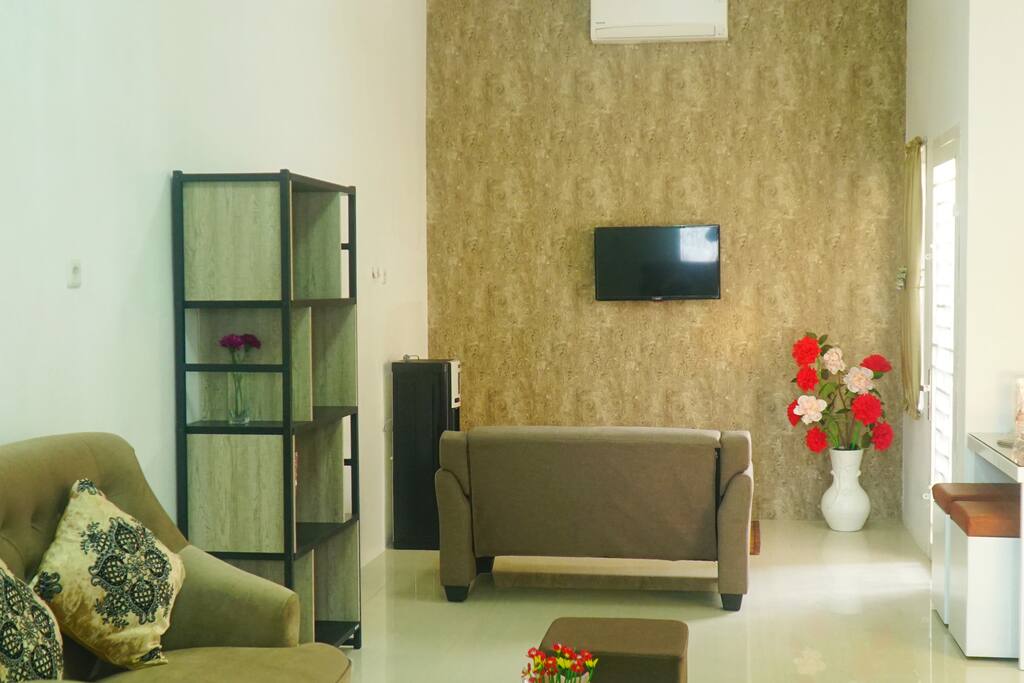 Ruang keluarga yang nyaman dilengkapi dengan sofa 2 seat, meja, televisi, AC, kipas angin, karpet, dan standing dispenser.