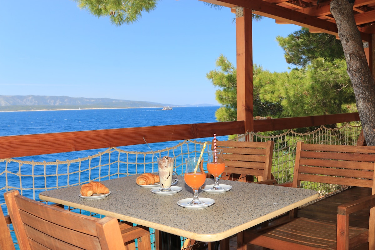 A shaded terrace is featured with a wooden table and four chairs, overlooking the sea. Two vibrant orange drinks are positioned alongside a dessert. Croissants are placed on white plates, with the blue water stretching out in the background under a clear sky.