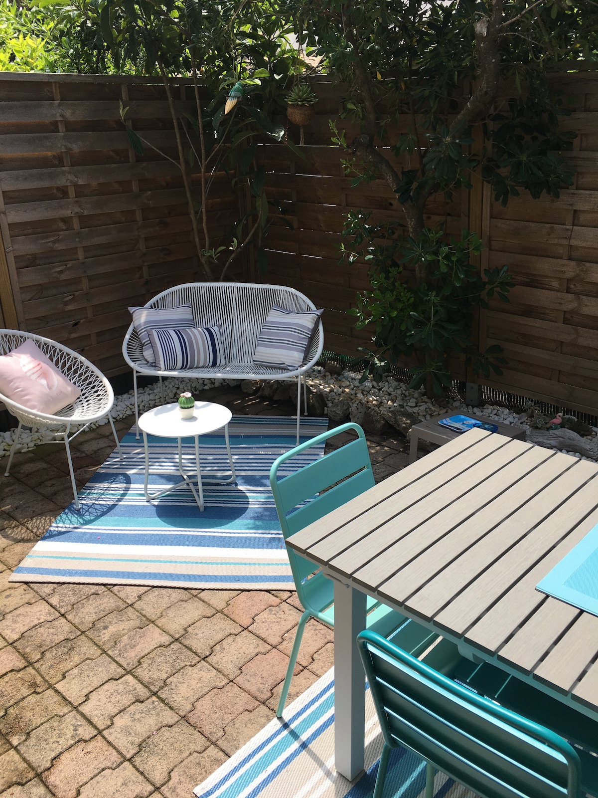 A sunlit terrace features a light blue and white striped rug. Two woven chairs with cushions and a small white table provide a cozy seating area surrounded by greenery. A dining table with matching chairs is also present, inviting relaxation outdoors.