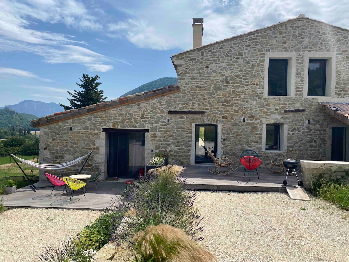 The exterior of a renovated stone barn showcases a spacious terrace with colorful chairs and a hammock. A barbecue grill and natural landscaping add to the inviting outdoor setting. The backdrop features rolling hills and clear skies, enhancing the serene countryside atmosphere.